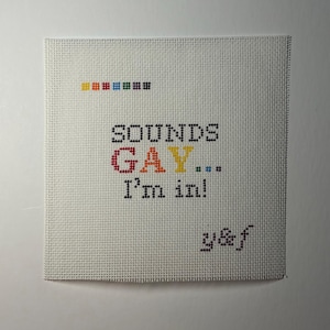 May include: A square white needlepoint canvas featuring the black cross-stitched text "SOUNDS GAY... I'm in!" with the word "GAY" in multicolored letters. A small row of colored squares sits above the text, and the letters "y&f" appear in the bottom right corner.