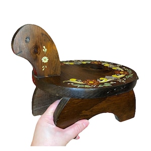 May include: A dark brown wooden stool shaped like a turtle, featuring a circular seat with a hand-painted floral wreath design in yellow, orange, and green. The stool has a curved head piece with a small painted flower and four sturdy legs.