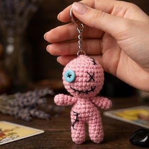 Könnte beinhalten: Ein rosa gehäkelter Puppen-Schlüsselanhänger mit einem blauen Knopfauge und schwarzen Nahtdetails. Die Puppe hat einen genähten Mund und kreuzgestickte Augen. Der Schlüsselanhänger ist an einem silbernen Metallring befestigt.
