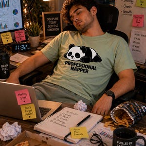 Może przedstawiać: Jasnozielona koszulka z czarno-białą grafiką pandy i napisem "PROFESSIONAL NAPPER". Na zdjęciu osoba śpiąca przy biurku z laptopem, notatnikiem i kubkiem do kawy. Na kubku napis "FUELLED BY CAFFEINE & DEADLINES."