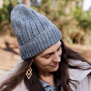 May include: A woman wearing a gray knitted beanie with a cable knit pattern and wooden geometric earrings. She is wearing a white jacket and has long brown hair.