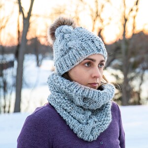 May include: A grey knitted hat with a fluffy pom pom and a matching grey knitted scarf. The hat and scarf are made with a chunky yarn and have a cable knit pattern.