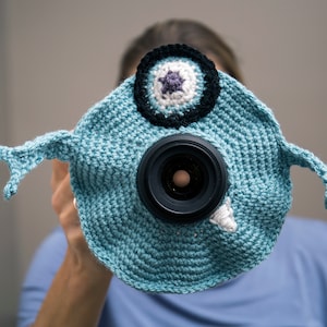 May include: A light blue crocheted camera cover shaped like a cartoon monster, with a large black lens as the eye. The cover has a white tooth and a circular design with black, white, and purple accents. The monster has two small arms.