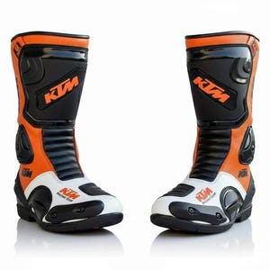 Puede incluir: Par de botas de moto naranjas, negras y blancas. Las botas tienen el logotipo de KTM en naranja y negro. Las botas están hechas de cuero y tienen paneles protectores. Las botas están diseñadas para carreras y tienen un aspecto elegante y moderno.