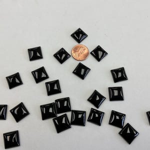 May include: A collection of glossy, black, square beads with rounded edges are scattered on a white surface. A penny is included for scale. The beads have a reflective surface and are likely for crafting or jewellery.