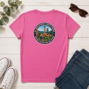 May include: A pink t-shirt featuring a circular graphic of a historical society and museum. The graphic includes a building, water tower, and text that reads "Keeping the Past for the Future".
