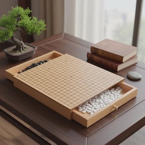 Könnte beinhalten: Ein hölzernes Go-Brettspiel-Set mit schwarzen und weißen Spielsteinen. Das Brett ist geöffnet und zeigt eine Schublade voller weißer Steine. Ein kleiner Bonsai-Baum und Bücher befinden sich im Hintergrund.