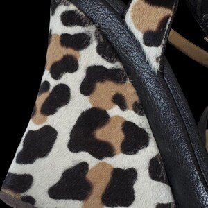 May include: Close-up of a wedge heel shoe with a black leather strap and a leopard print design. The print features black, brown, and white spots on a textured surface. The shoe has a tan strap.