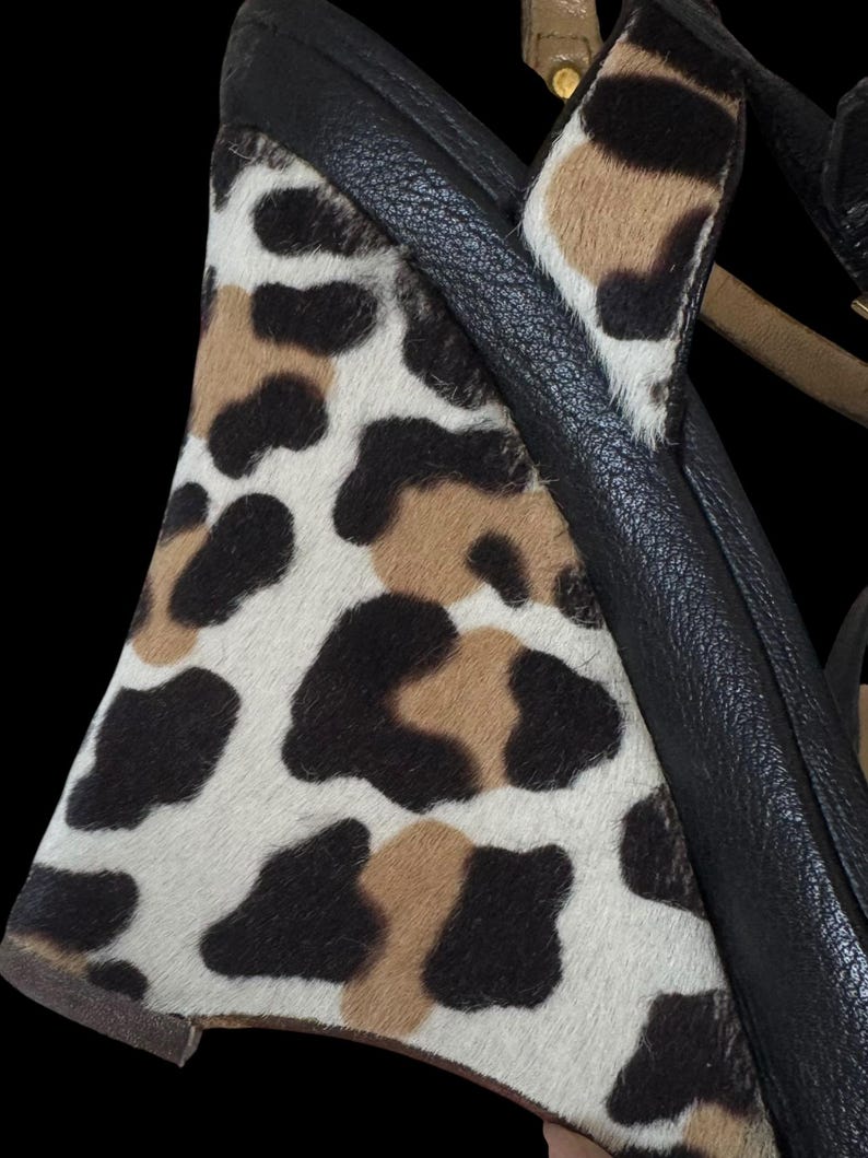 May include: Close-up of a wedge heel shoe with a black leather strap and a leopard print design. The print features black, brown, and white spots on a textured surface. The shoe has a tan strap.