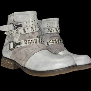 May include: A pair of light gray ankle boots with silver-tone studs and buckles. The boots have a distressed leather appearance, with decorative straps and a patterned fabric detail. The soles are brown with small studs.