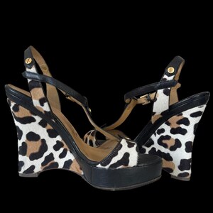 May include: A pair of wedge sandals with a black platform and straps. The wedge heels feature a leopard print pattern in black, brown, and white. The sandals have gold-tone hardware.