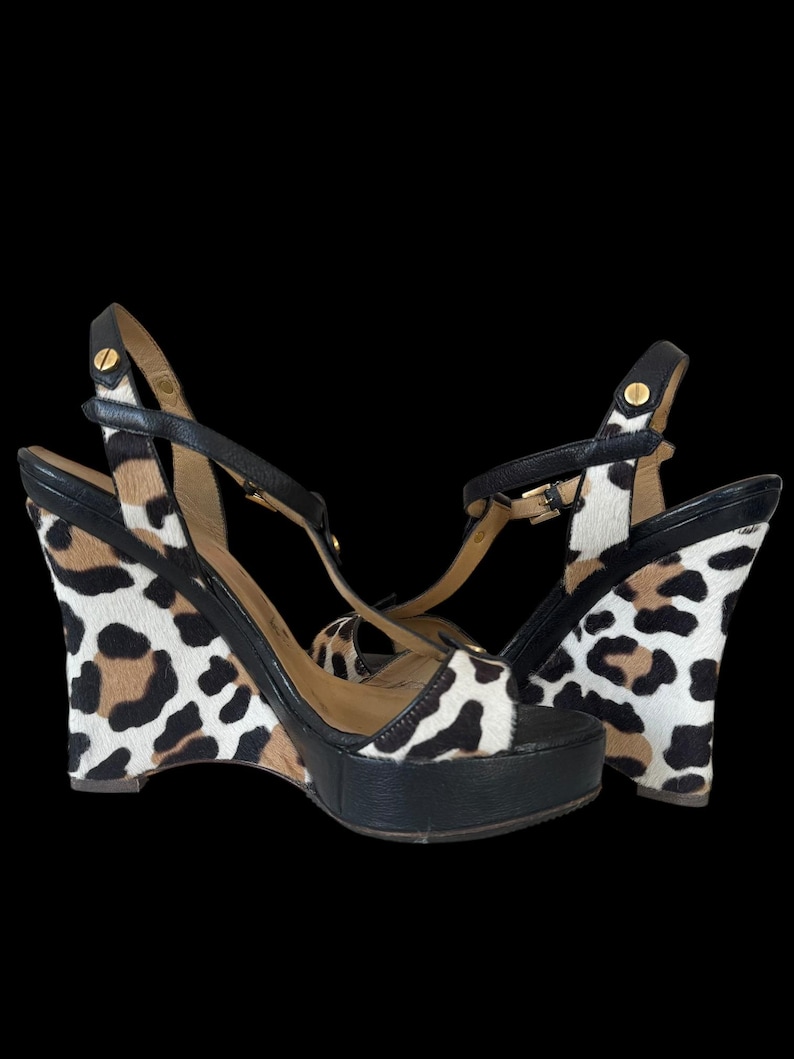 May include: A pair of wedge sandals with a black platform and straps. The wedge heels feature a leopard print pattern in black, brown, and white. The sandals have gold-tone hardware.