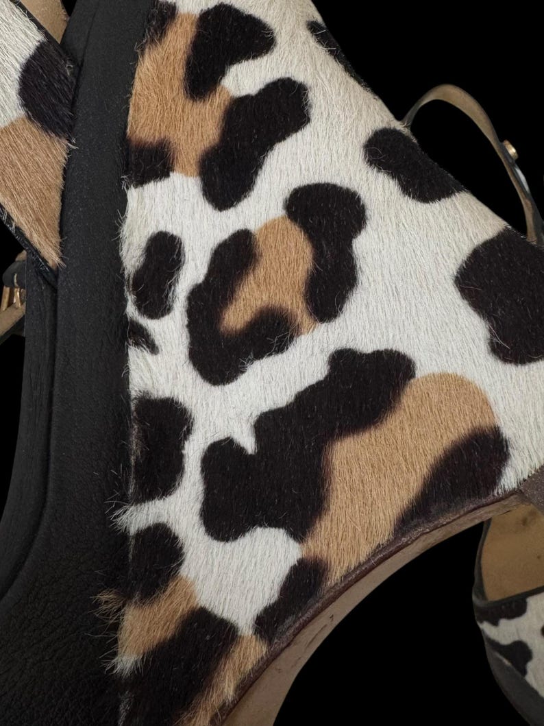 May include: Close-up of a pair of high-heeled shoes with a black leather base and a spotted pattern in white, brown, and black. The shoes have a thin ankle strap and a high heel.