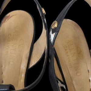 May include: Close-up of two black leather Valentino Garavani sandals. The sandals feature thin straps, gold-colored hardware, and a leopard print accent. The brand name is visible on the insole.