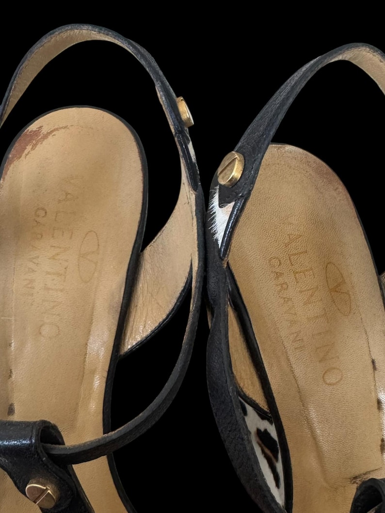May include: Close-up of two black leather Valentino Garavani sandals. The sandals feature thin straps, gold-colored hardware, and a leopard print accent. The brand name is visible on the insole.