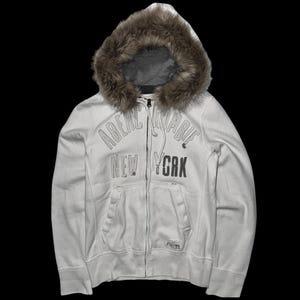 May include: A white zip-up hoodie featuring a brown faux fur-lined hood and long sleeves. The front displays the text Abercrombie New York in large, embroidered lettering. It includes two front pockets and a full-length metal zipper closure.