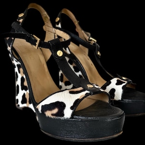 May include: A pair of platform wedge sandals with a black and white animal print design. The sandals have black straps and soles, with gold-colored hardware. The wedge heel is approximately 5 inches high.