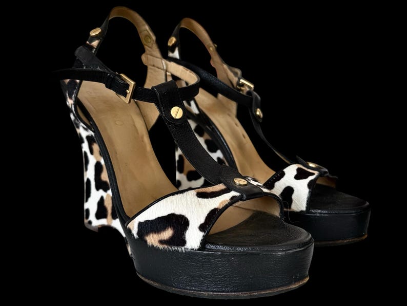 May include: A pair of platform wedge sandals with a black and white animal print design. The sandals have black straps and soles, with gold-colored hardware. The wedge heel is approximately 5 inches high.