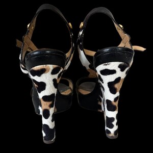 May include: Pair of high-heeled sandals with a black leather strap and a cowhide pattern on the heels. The heels are approximately 4 inches tall. The sandals have a buckle closure and gold-colored hardware.