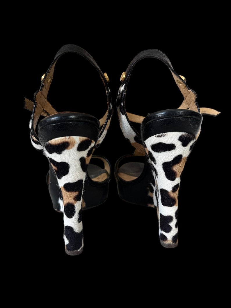 May include: Pair of high-heeled sandals with a black leather strap and a cowhide pattern on the heels. The heels are approximately 4 inches tall. The sandals have a buckle closure and gold-colored hardware.