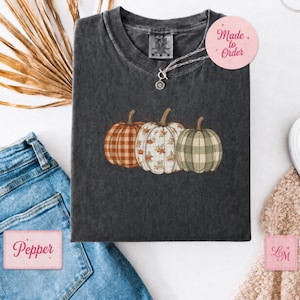 May include: A charcoal grey short-sleeve t-shirt featuring a graphic of three pumpkins. The left pumpkin has an orange and white plaid pattern, the centre pumpkin has a white floral print, and the right pumpkin has a green and white plaid pattern.