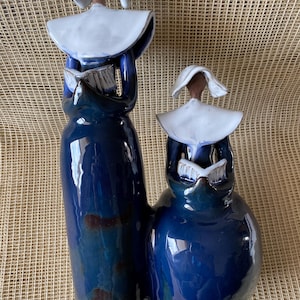 Puede incluir: Dos esculturas de cerámica de figuras con túnicas azules y cofias blancas, cada una sosteniendo un libro. Las figuras están conectadas en la base, con un acabado brillante y un toque de marrón. El fondo es una tela tejida beige.