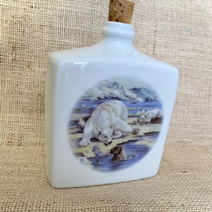 Può includere: Bottiglia in ceramica bianca con tappo in sughero. La bottiglia presenta un'illustrazione circolare di orsi polari in un paesaggio innevato. Un orso è vicino all'acqua e un altro è nell'acqua. La bottiglia ha una forma quadrata.