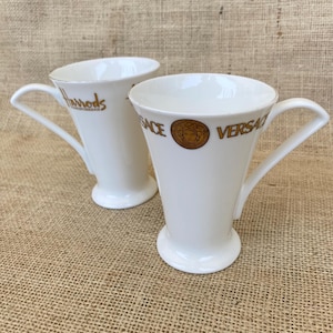 May include: Two white porcelain mugs with flared rims and pedestal bases. One mug features the gold text Harrods Knightsbridge, while the other displays the gold text Versace and a circular gold Medusa head emblem on the front exterior surface.