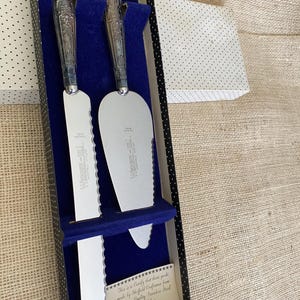 Può includere: Un set di coltello e paletta per torte argentati in una scatola a pois bianchi e neri con interno blu. I manici hanno un design ornato. Il set include un coltello con bordo seghettato e un server. Il testo sulla scatola recita "WERBER-HILL SHEFFIELD CUTLERY".