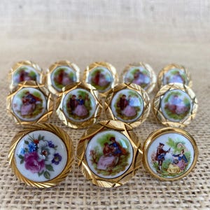 Può includere: Una collezione di undici bottoni rotondi con bordi in metallo dorato caratterizzati da un motivo testurizzato a raggiera. Dieci bottoni mostrano scene dipinte in miniatura di figure all'aperto, mentre un bottone presenta un motivo floreale.