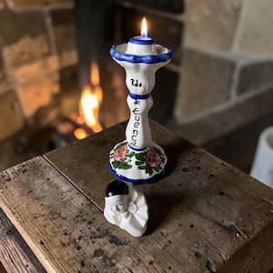 May include: A white ceramic candlestick holder with blue trim, hand-painted floral accents, and the text Rdo. de CUENCA, holding a lit candle. Beside it sits a small white ceramic figurine of a person with a black cap and a ruffled collar.