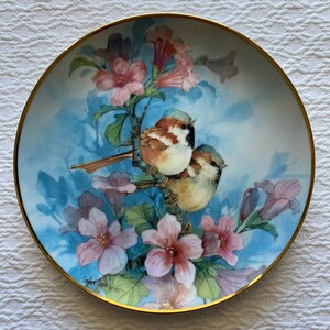 Könnte beinhalten: Dekorative Platte mit goldenem Rand, die zwei kleine Vögel auf einem blühenden Ast mit rosa Blüten darstellt. Der Hintergrund ist hellblau. Das Kunstwerk ist signiert.