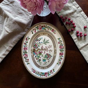 May include: An oval ceramic serving platter featuring a cream-colored base with a central illustration of a flowering tree branch in shades of pink, blue, and green, surrounded by a decorative brown border and a floral rim pattern of pink roses.