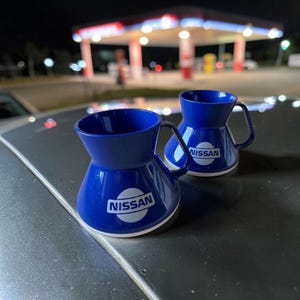 Könnte beinhalten: Zwei blaue Kunststoffbecher mit ausgestelltem, breitem Bodendesign und einem einzelnen Henkel. Jeder Becher ist auf der Vorderseite mit dem weißen Nissan-Logo bedruckt. Die Becher haben eine glatte, glänzende Oberfläche und einen weißen Bodenrand.