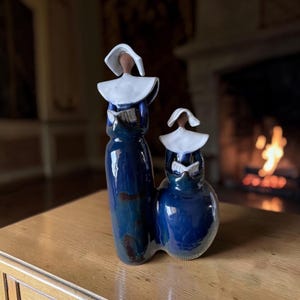 May include: A ceramic sculpture featuring two figures in dark blue robes with white head coverings and collars. The taller figure stands on the left, while a shorter figure stands on the right. Both figures hold open books in their hands.