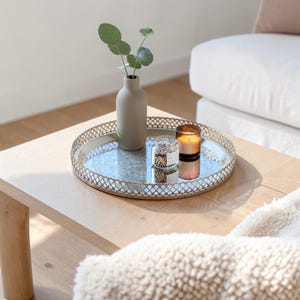Könnte beinhalten: Ein rundes Spiegeltablett mit einem dekorativen metallischen Gitterrand. Auf dem Tablett stehen eine matte, beige Keramikvase mit grünen Eukalyptuszweigen, ein kleines Glas mit Streichhölzern und ein Glasbehälter mit einer brennenden Kerze.
