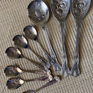 Könnte beinhalten: Eine Sammlung von Vintage-Servierlöffeln und einer Gabel aus Silber. Die Löffel und die Gabel weisen aufwendige Blumen- und Fruchtdesigns auf den Laffen und Griffen auf. Die Utensilien sind fächerförmig auf einer strukturierten, neutralfarbenen Oberfläche angeordnet.