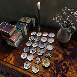 May include: A collection of twenty-one miniature ceramic plates arranged on a wooden surface. The plates feature various decorative patterns, including floral motifs, landscape scenes, and solid colors, with some having scalloped or gold-toned edges.