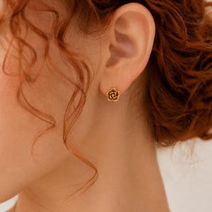 Könnte beinhalten: Ein kleiner goldener Ohrstecker in Form einer blühenden Rose. Der Ohrring zeigt ein detailliertes, mehrschichtiges Blütenblatt-Design mit einem zentralen Punkt und ist in einer polierten, goldfarbenen Metalloberfläche gefertigt.