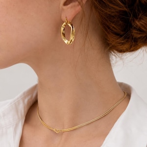 Könnte beinhalten: Ein goldfarbener, gedrehter Creolen-Ohrring und eine dünne, goldfarbene Halskette mit einem kleinen Knoten-Detail in der Mitte. Das Schmuckset wird zu einem weißen Hemd mit Kragen getragen und zeigt ein poliertes Metall-Finish.