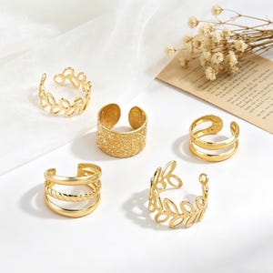 May include: Five gold-colored adjustable rings. Designs include a leaf pattern, textured band, double band, and a twisted rope design. The rings are displayed on a white surface with dried flowers and a piece of paper in the background.