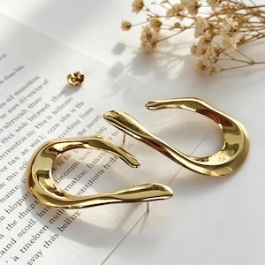 May include: A pair of gold-colored, abstract-shaped earrings with a post backing. The earrings have a smooth, reflective surface and a unique, curved design. A single earring post is visible.
