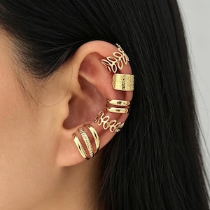 May include: A collection of gold-colored ear cuffs in various designs. The ear cuffs include a leaf design, a textured rectangle, double bands, and openwork patterns. The jewelry is worn on the ear, showcasing the different styles and textures.