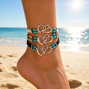 May include: Three anklets with silver heart charms and turquoise beads are displayed on an ankle at the beach. The top anklet has white and black string, the middle one has colorful beads, and the bottom one is black.
