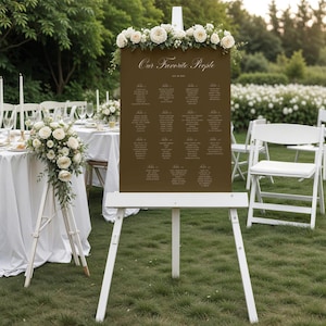 May include: A dark brown rectangular wedding seating chart sign titled Our Favorite People, displayed on a white wooden easel. The sign is decorated with a floral arrangement of white roses and greenery at the top and lists guest names.