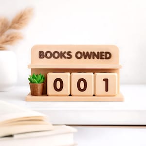 Può includere: Un espositore in legno beige con la scritta "BOOKS OWNED" e tre blocchi numerati che mostrano "001". Una piccola pianta grassa in vaso è a sinistra. Il tutto è su una superficie bianca.