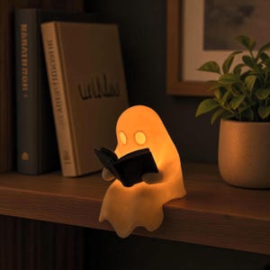 Può includere: Una piccola lampada in silicone a forma di fantasma color bianco sporco. Il fantasma ha due occhi ovali ed è seduto sul bordo di uno scaffale, tenendo tra le mani un piccolo libro nero aperto. La lampada emette una luce arancione calda e soffusa.