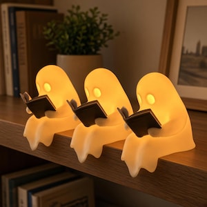 Op de afbeelding: Drie kleine, oplichtende, gebroken witte lampen in de vorm van een geest, elk zittend op een rand en een klein, donker, open boek vasthoudend. De lampen verspreiden een warm, zacht licht dat hun gladde vormen en oogvormen benadrukt.
