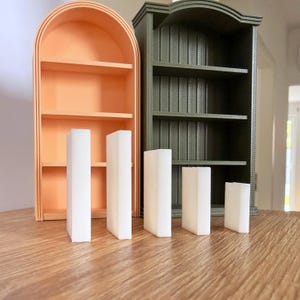 Op de afbeelding: Twee miniatuur boekenplanken, één perzik van kleur met een gewelfde bovenkant en de andere donkergroen. Vier witte rechthoekige blokken staan voor de planken. De planken hebben meerdere niveaus voor het presenteren van kleine items.