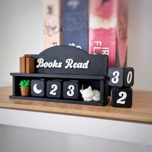 Könnte beinhalten: Ein schwarzes Holzregal "Books Read" mit weißen Buchstaben. Es zeigt eine kleine Topfpflanze, einen Halbmond, die Zahlen 23 und eine weiße Katzenfigur. Zwei schwarze Würfel zeigen die Zahlen 30 und 2. Bücher sind im Hintergrund.